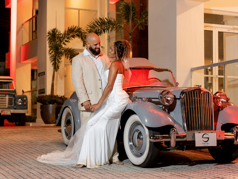 Bride and groom in elegant attire standing beside a vintage car at entrance of S Hotel Montego Bay at night with warm lighting and palm trees creating a romantic setting.