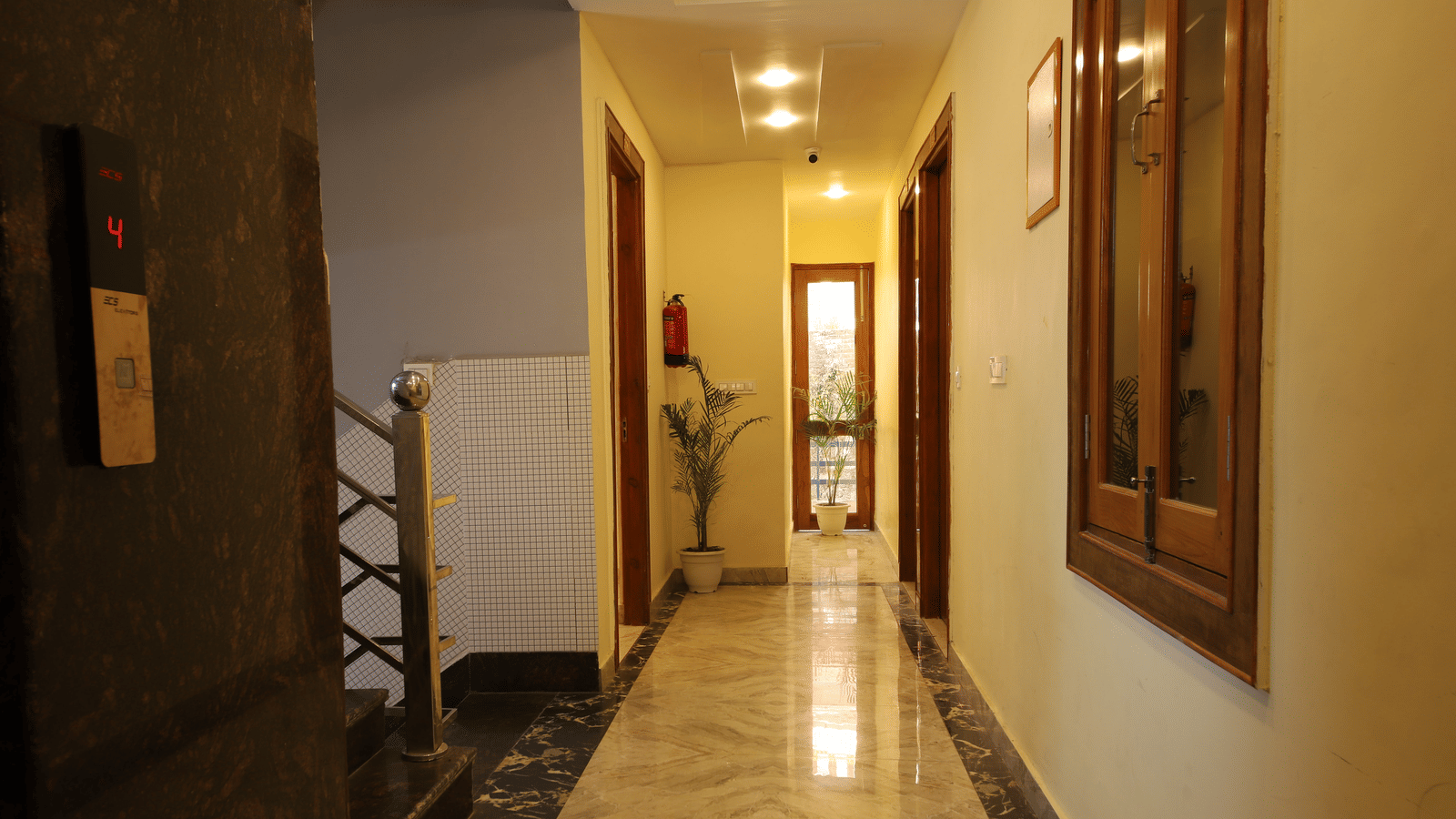 A view of a narrow hotel hallway with marble flooring, wooden doors, a mirror, and dim lighting | Perfectstayz Value Hills