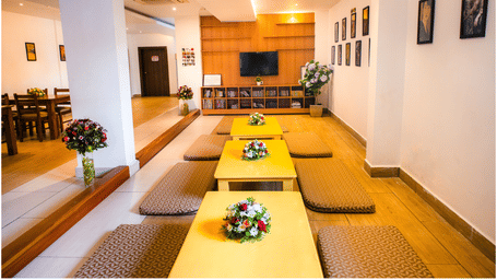 Japanese-style floor seating with low tables and cushions beside book shelves and a television in a wooden shelf wall at Ajito, Ilara Hotels & Spa, Tada.