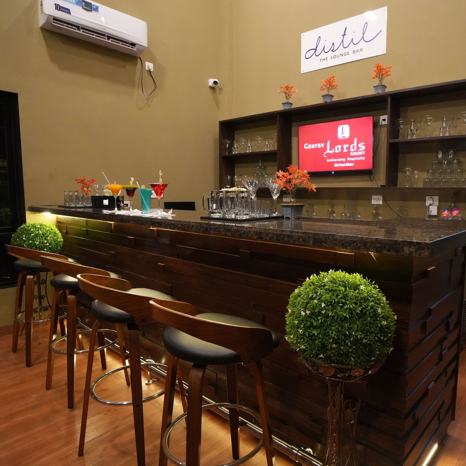Modern indoor lounge bar with a sleek wooden counter, stylish high chairs, decorative plants, glass shelves, and a variety of colourful cocktails on display - Gaurav Lords Resort Shrivardhan