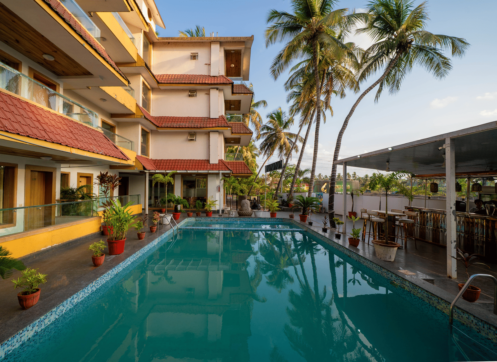 A sunny outdoor swimming pool is located within the courtyard surrounded by palm trees and buildings at Perfectstayz Premium Goa Continental.