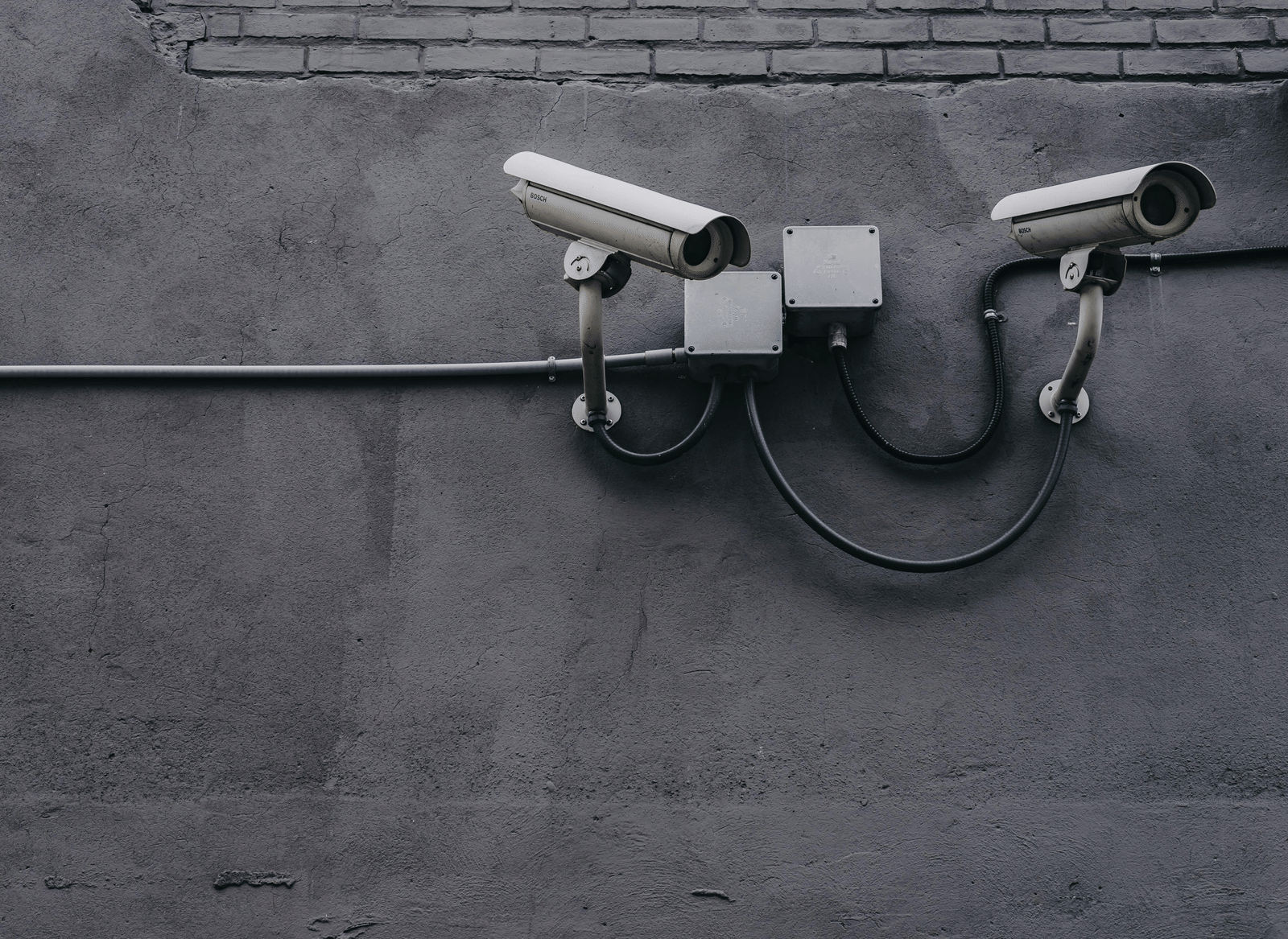 Two surveillance cameras mounted on a grey concrete or brick wall, with visible wiring.