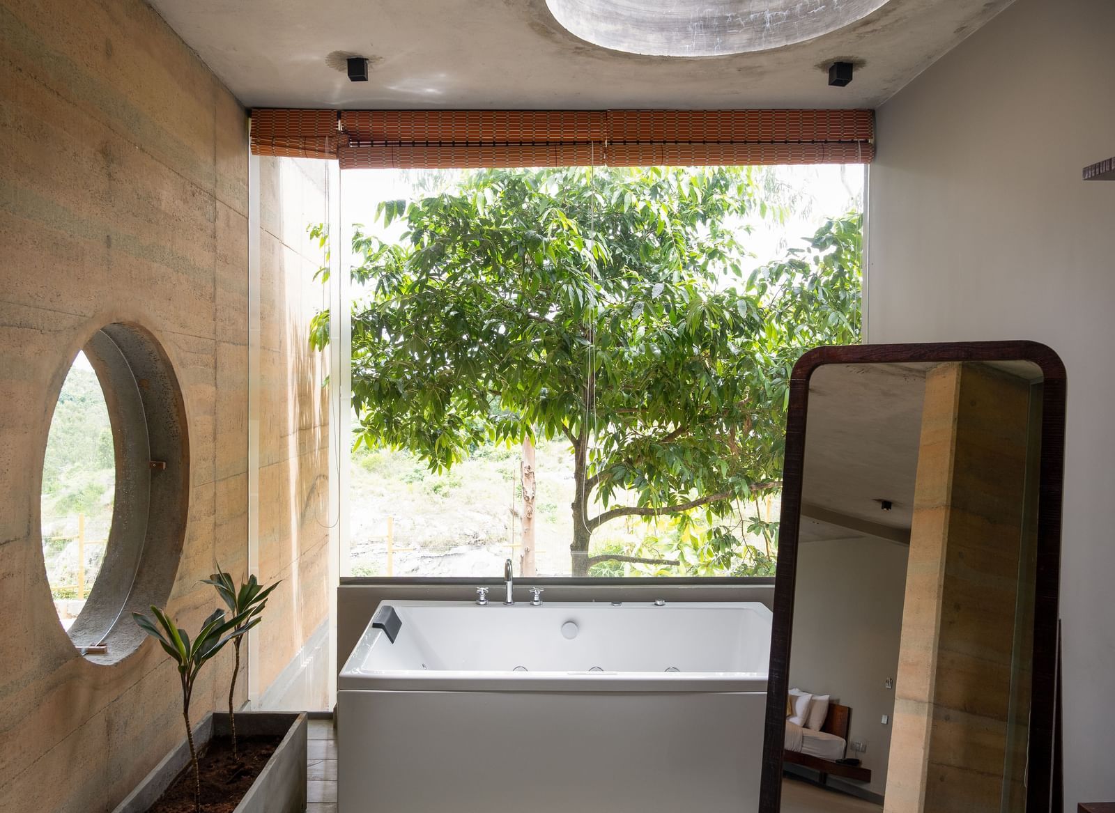 A bathroom featuring a large round mirror, a vanity area, and a window overlooking lush trees | Gaia Studio | RD's Nature Retreat, Bangalore