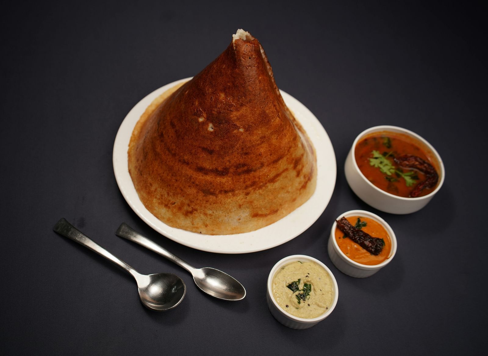 A golden dosa on a white plate, accompanied by three small bowls of colourful chutneys and sambar, with two spoons resting beside it.
