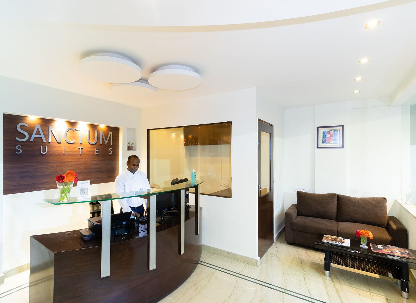 The reception area at Sanctum Suites Domlur, Bangalore featuring a reception desk, a brown sofa, and a coffee table.