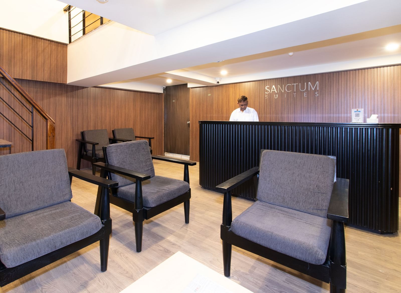 Reception area with grey armchairs, a reception desk, and a person in the background - Sanctum Suites Richmond Road, Bangalore
