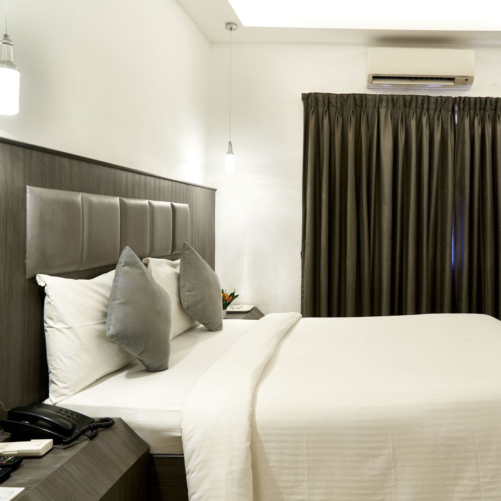 A modern hotel room at Hotel Mamallaa Heritage, featuring a bed with a dark upholstered headboard, white linens, and a flat-screen TV.