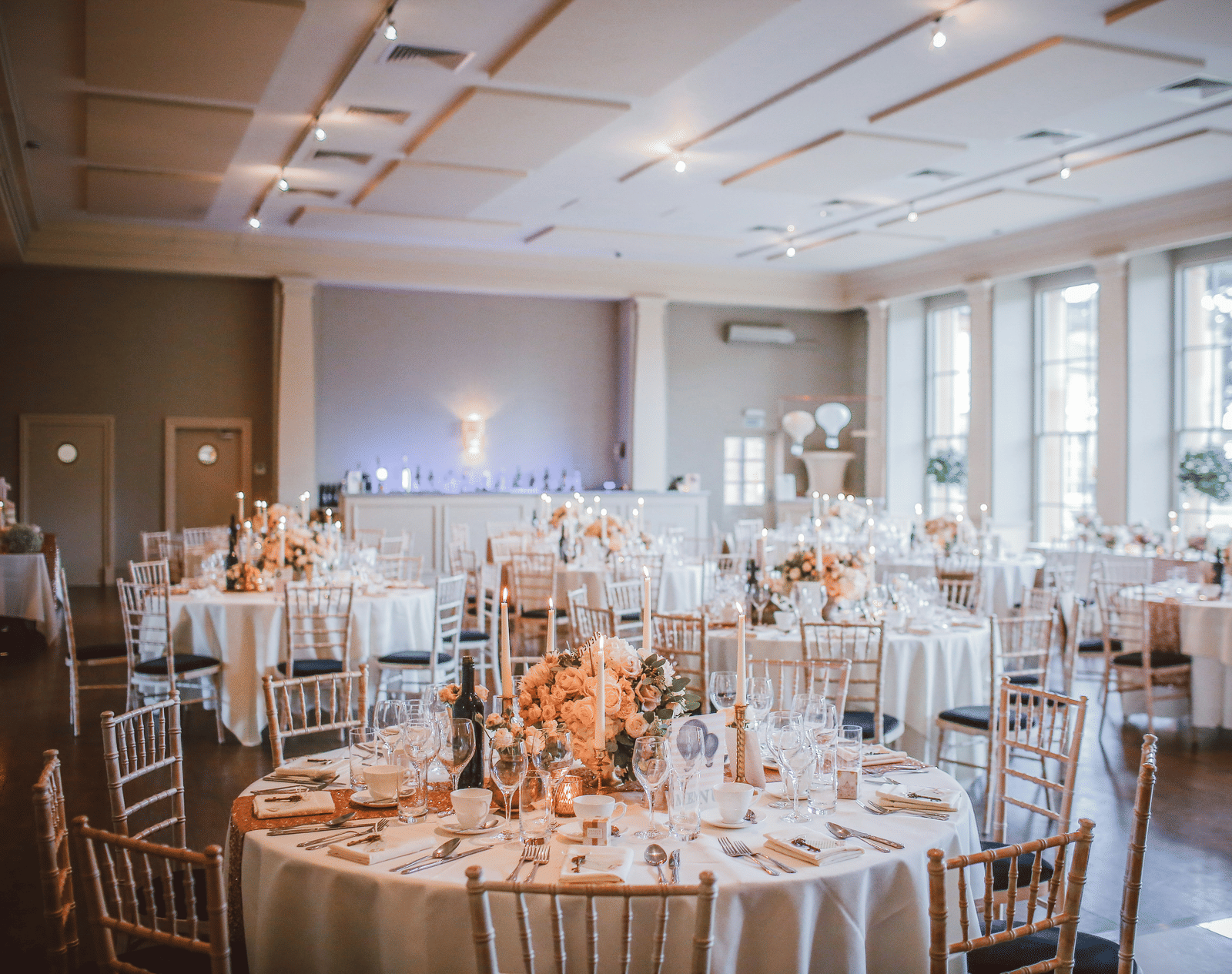 A wide view of an indoor wedding reception room with round tables, white linens, and large windows.