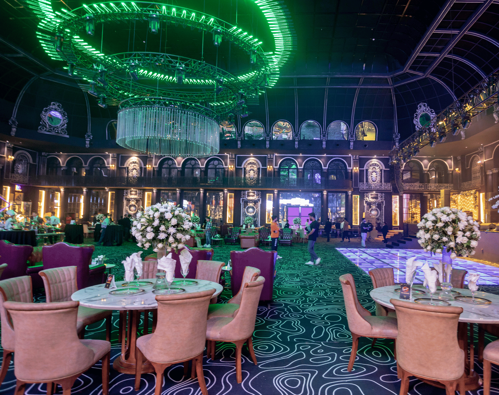 An opulent banquet hall featuring circular tables, pink chairs, and a large glowing green ceiling installation.