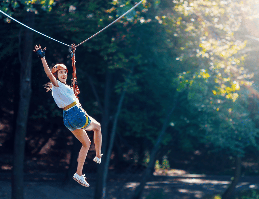 A young woman on a zipline with an excited expression on her face, on a bright sunny day with trees in the background.