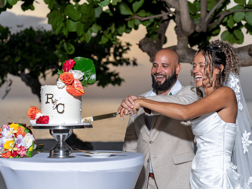 Bride and groom smiling while cutting a decorated wedding cake on a beachside table at sunset with tropical flowers and ocean view in the background.