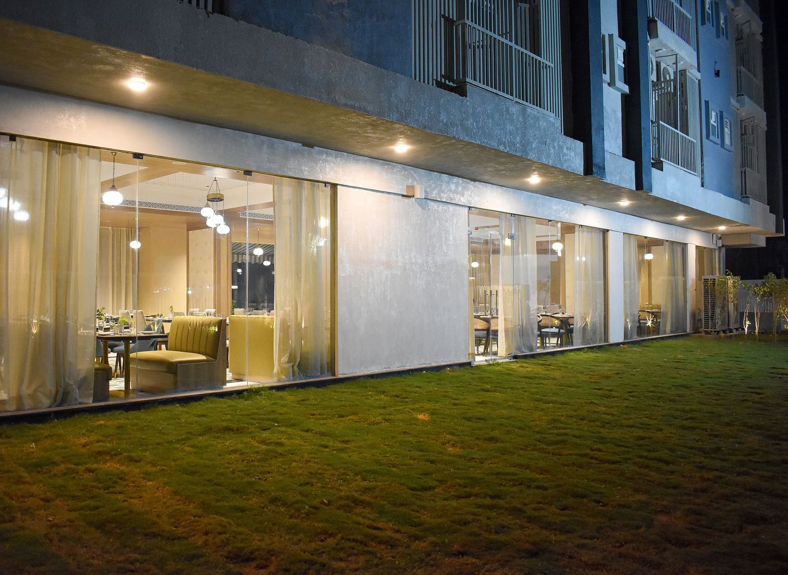 Exterior view of a building with a glass-walled restaurant, sheer curtains, hanging lights, and a grassy area in front - Urban Suites by BluSalzz, Jaipur