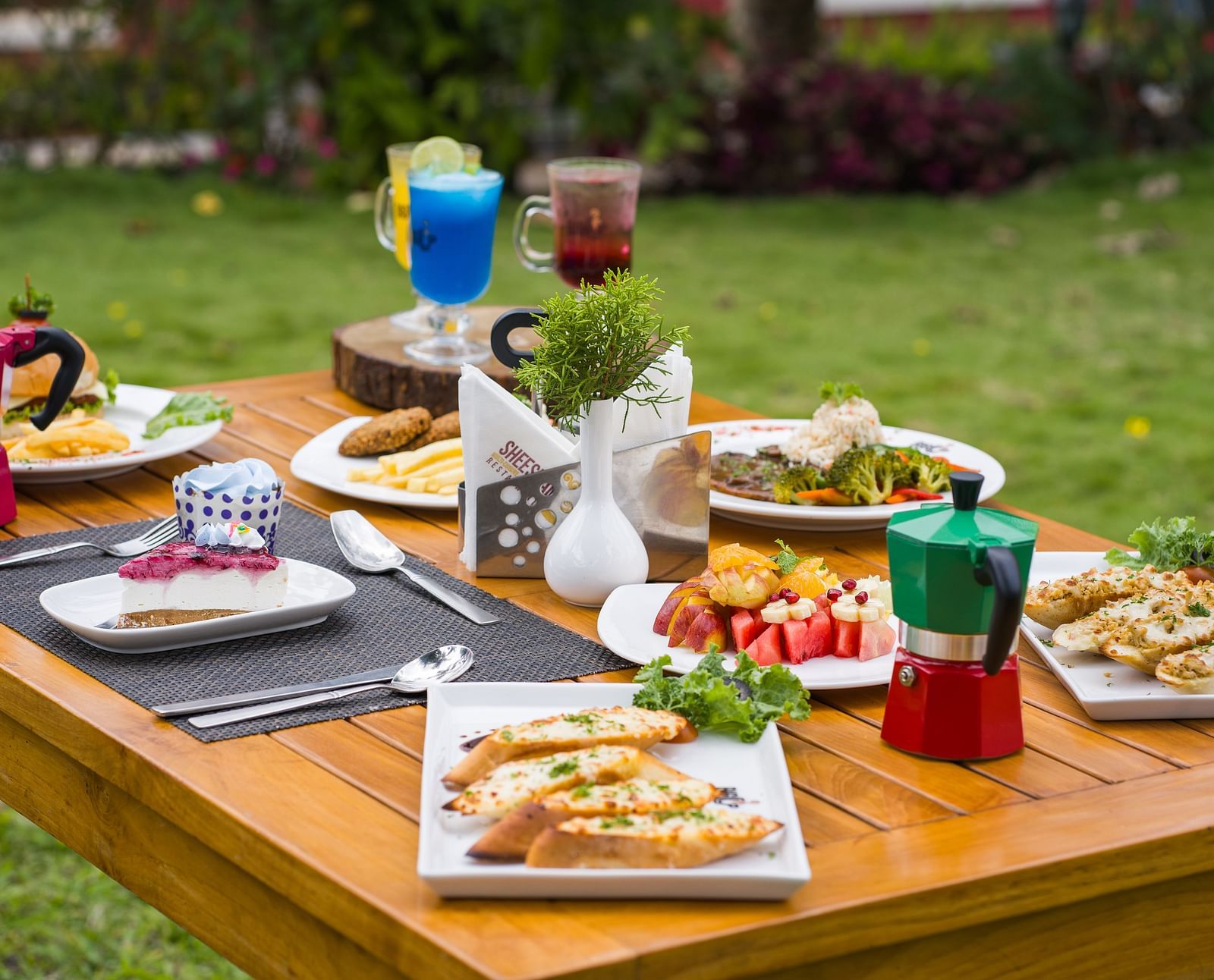 A wooden table is set outdoors with a variety of dishes and drinks, surrounded by a green lawn | Woodstock Resorts, Coorg