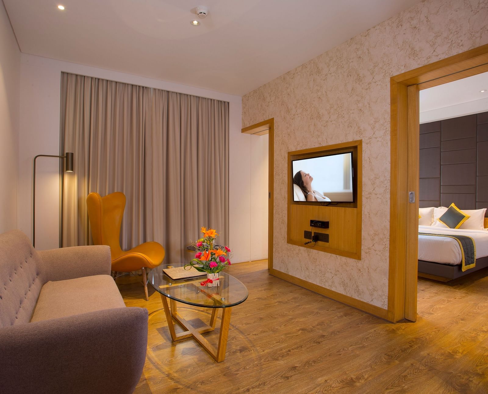 view of a living room with sofas and a small table, all facing a television mounted on the wall and a door leading to the bedroom on the right - Hotel Fountain Tree By TGI - JP Nagar, Bangalore