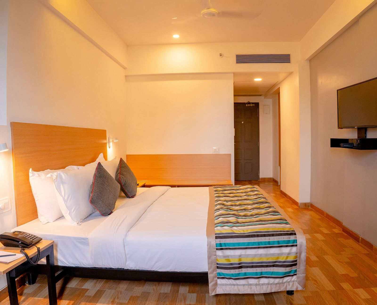 A cosy hotel room with a large bed, colourful throw and wooden furniture, including a desk with a TV above it - TGI Ameya Grand, Goa