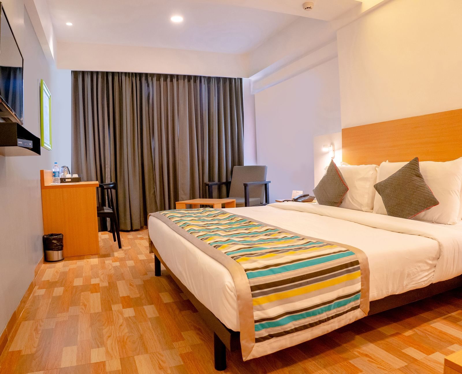 Another view of a cosy hotel room with a large bed, colourful throw and wooden furniture, including a desk with a TV above it - TGI Ameya Grand, Goa