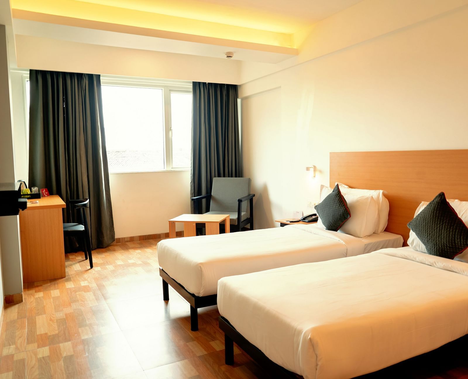 A bright hotel room with two single beds, a desk and a flat-screen TV, with a large window draped in gray curtains - TGI Ameya Grand, Goa