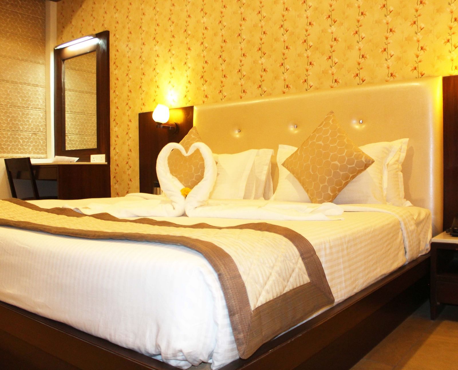 A warmly lit hotel room with a queen size bed, white bedding and a towel art swan on the bedspread - TGI Apple Inn, Jaipur 