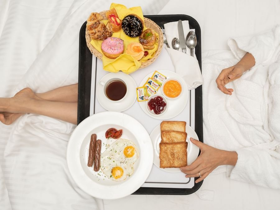 In-room breakfast tray with juice, fruits, and pastries placed beside a person on the bed at Heritage Village Resorts & Spa, Goa.