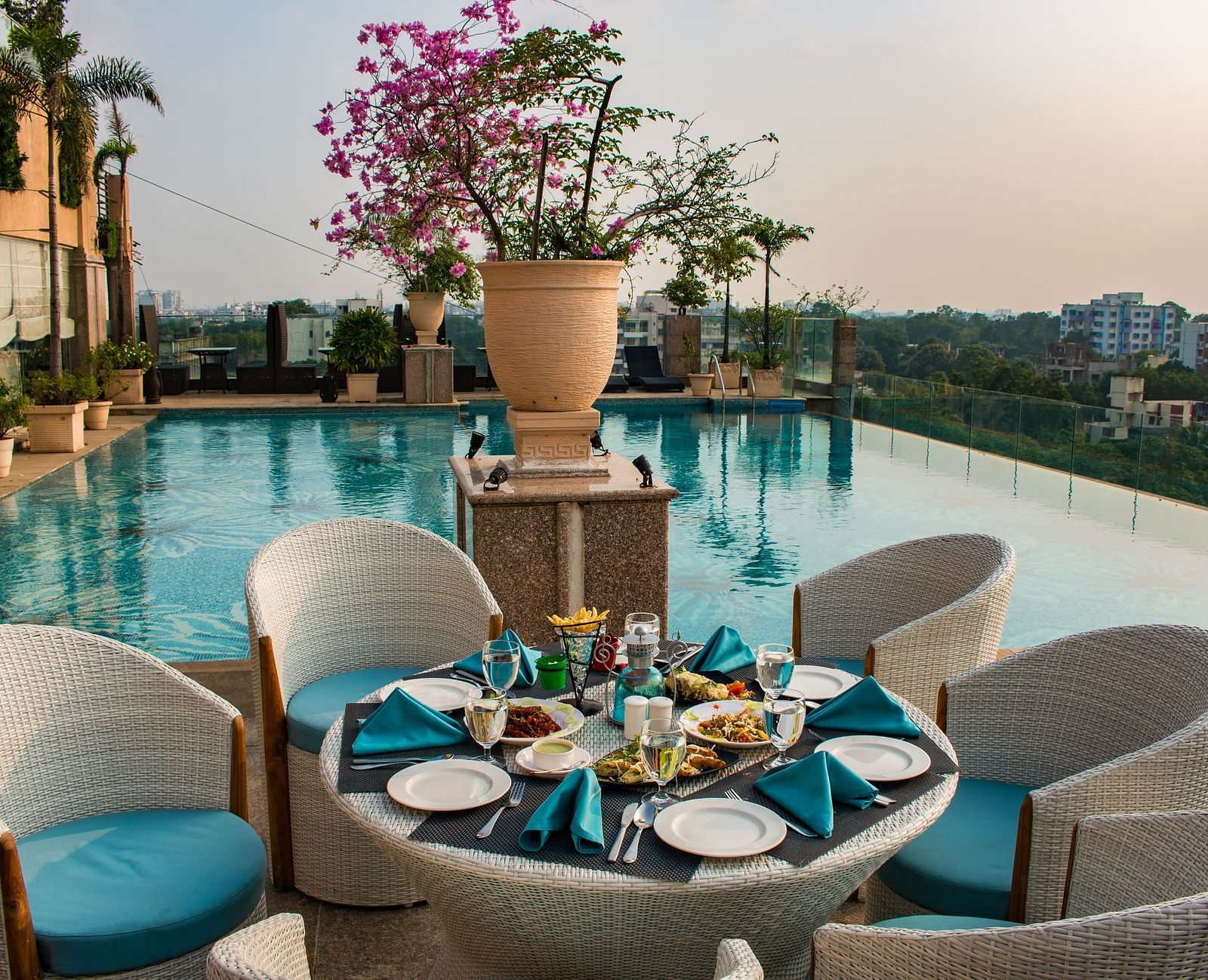 An outdoor dining at Hotel Ashok Nagpur featuring a round table with place settings for four, surrounded by cushioned chairs, and a swimming pool and cityscape in the background. 