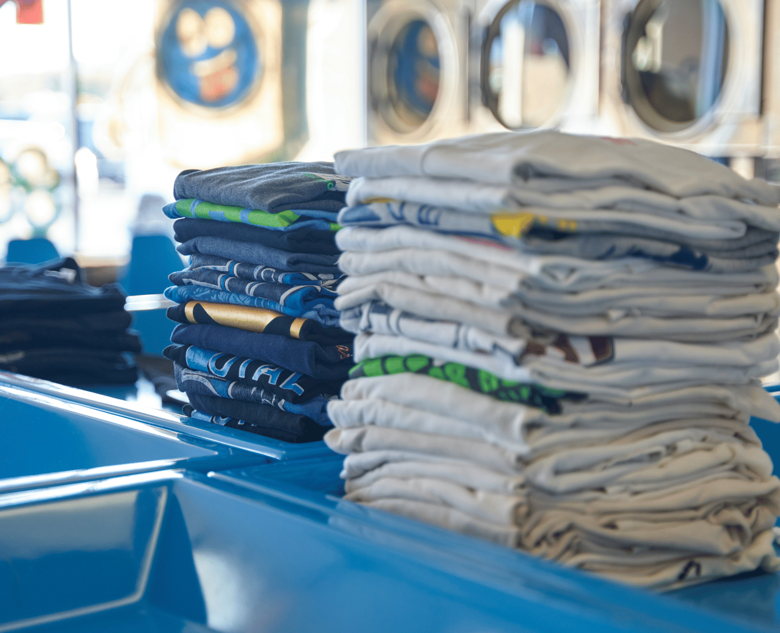 Multiple neat stacks of freshly folded laundry resting on a bright blue counter in a laundrette.