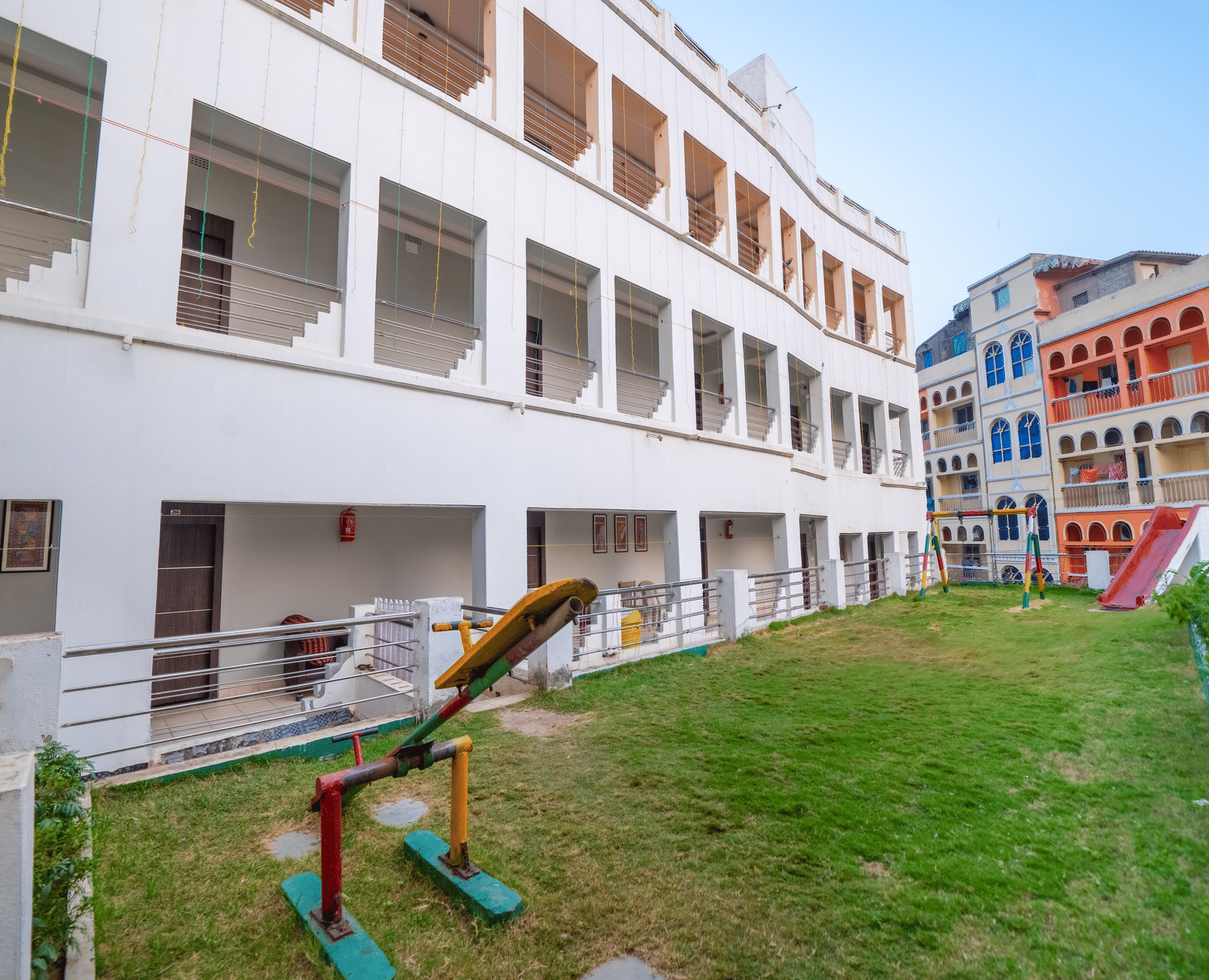 The exterior of a multi-storey, white modern hotel building surrounding a central courtyard with a green lawn and a small children's play area - Hotel Dolphin Puri