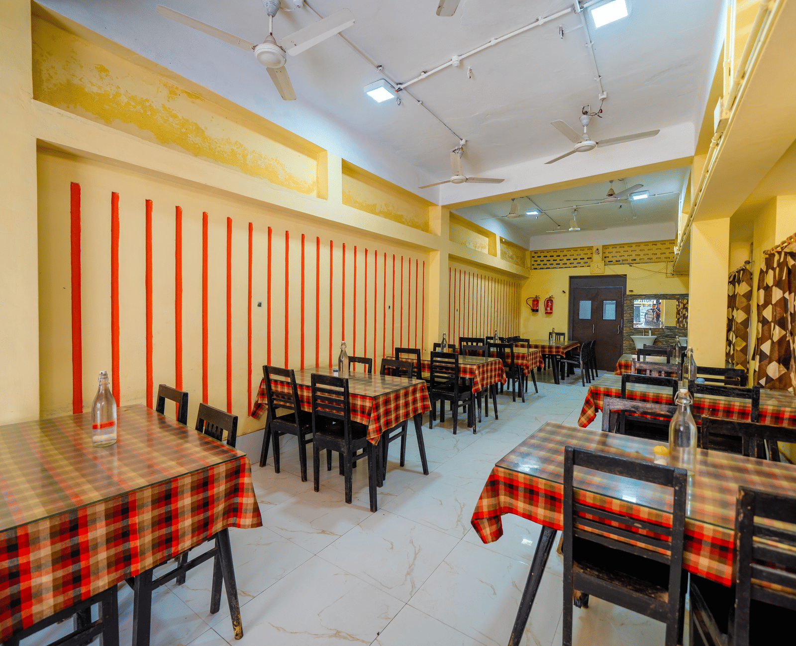 A brightly-lit, empty hotel dining hall with small tables covered in red and black checkered tablecloths and matching chairs - Hotel Dolphin Puri