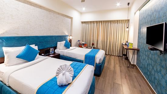 A corner view of the superior room at Icon Boutique Hotel by Bhagini, Domlur. The room features a twin bed, a flat-screen TV mounted on the wall, and a window with closed curtains, creating a peaceful and cozy atmosphere.