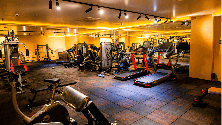 Fitness centre with an exercise bike, treadmills, a barbell rack and multi-gym stations under warm cove lighting at Ilara Hotels & Spa, Tada.