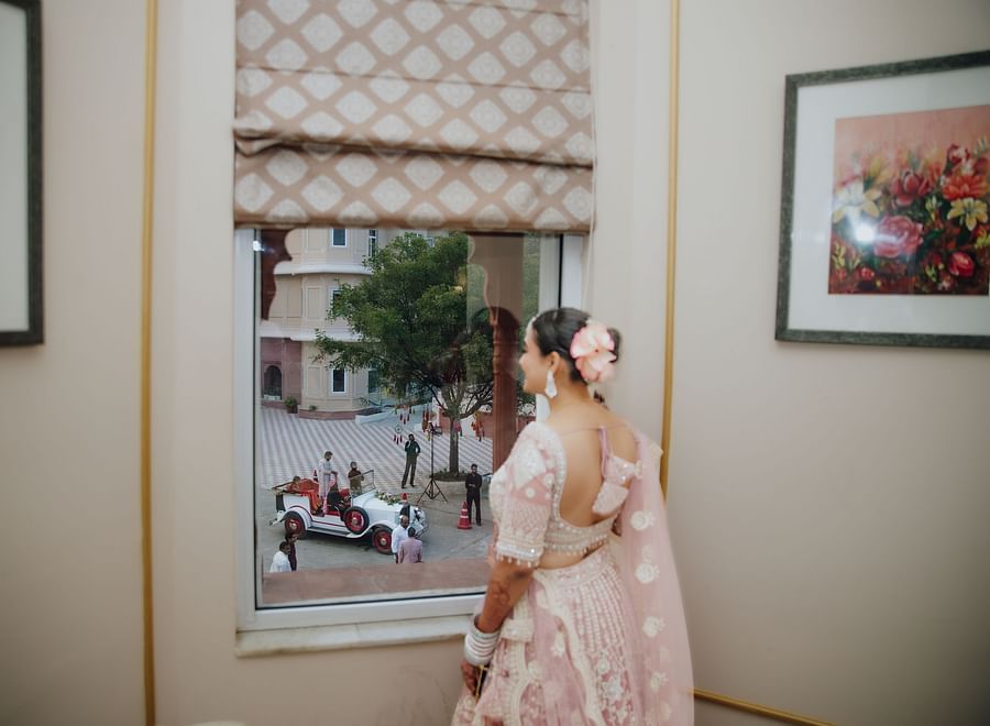 Bride standing by a window, smiling and looking outside at the groom who is waving from below.