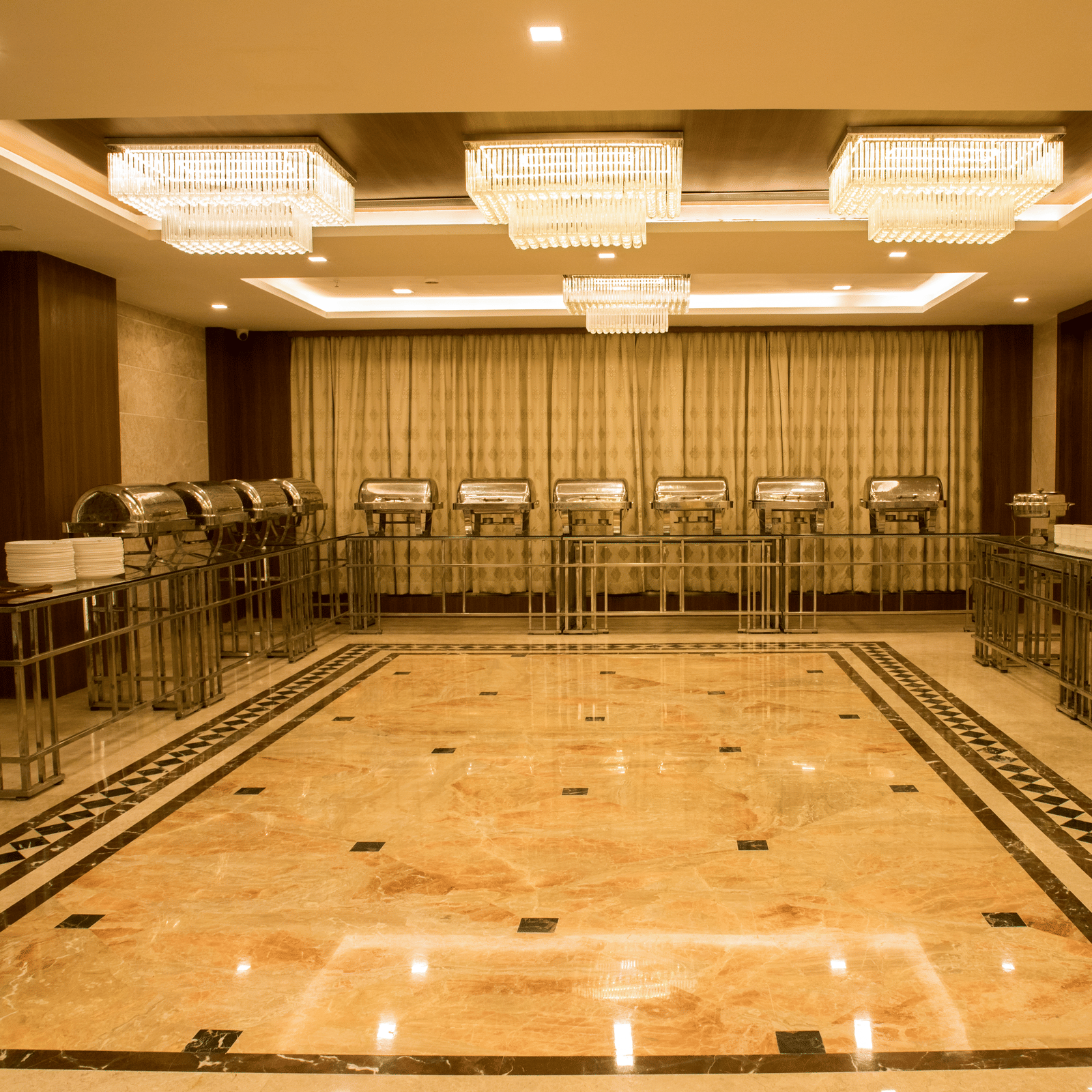 A brightly lit banquet hall with several chafing dishes lined at the far end of the hall at Benzz Park, Vellore.