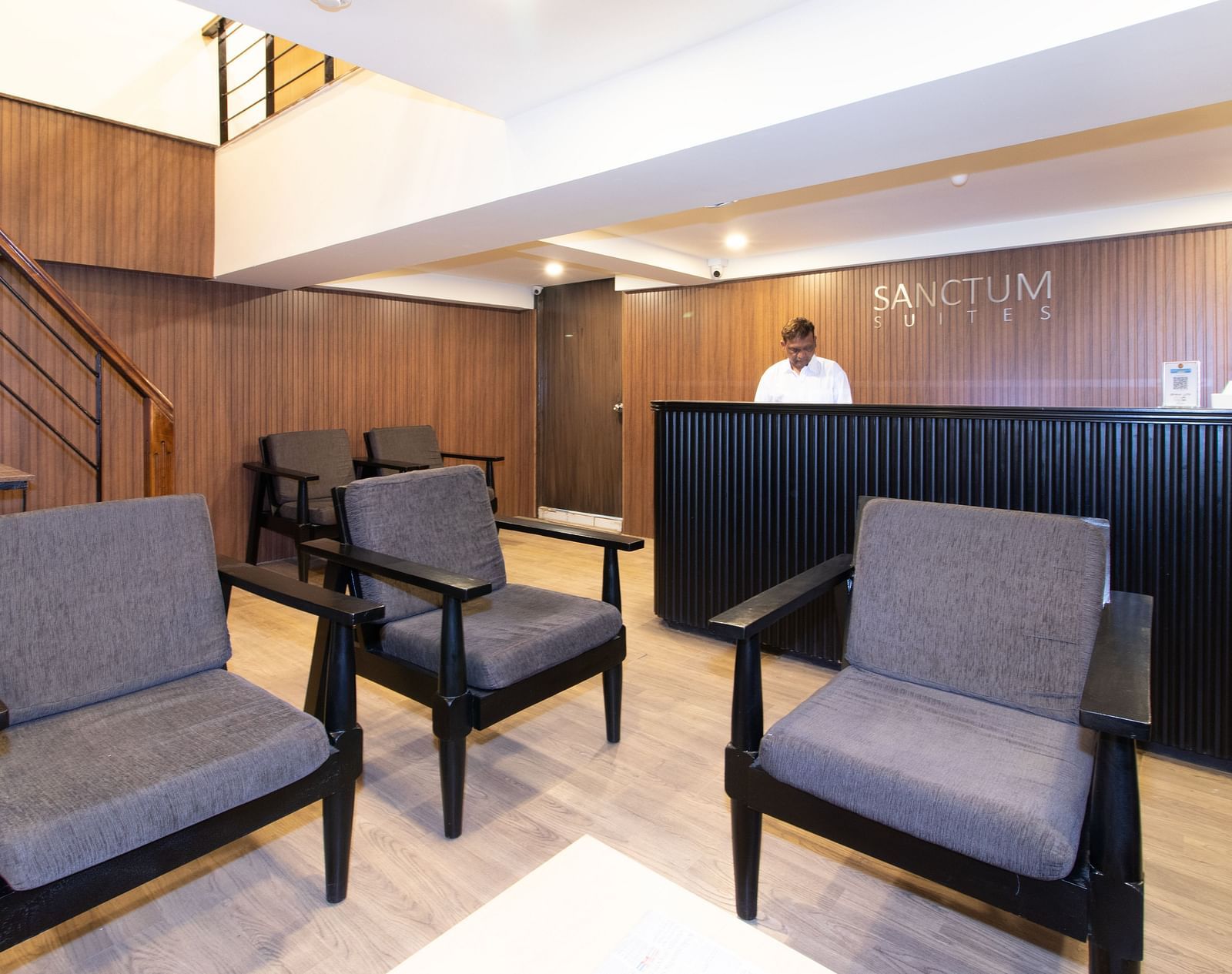 Reception area with grey armchairs, a reception desk, and a person in the background - Sanctum Suites Richmond Road, Bangalore