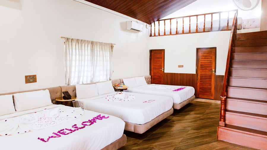 A spacious bedroom with multiple beds and a wooden staircase, featuring pink petals on the nearest bed spelling WELCOME | Vintage Trails By Ilara