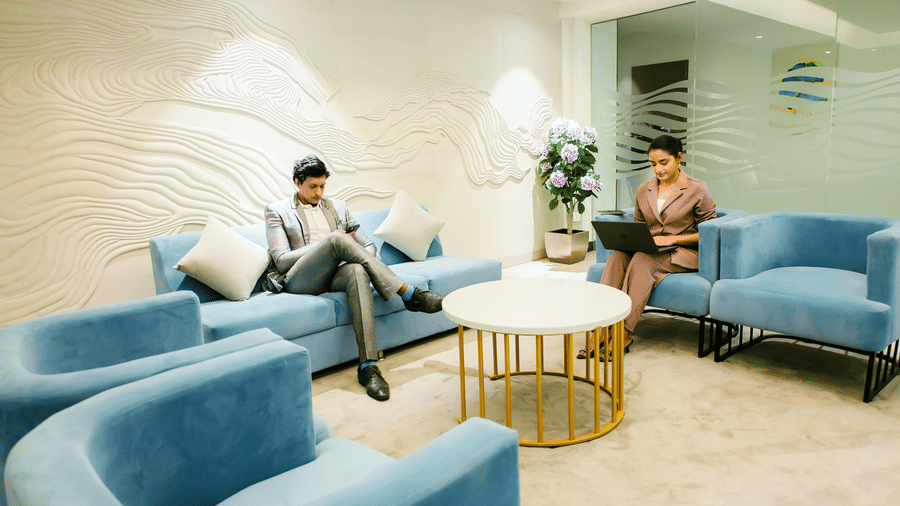 Guests using a laptop and phone on blue sofas around a coffee table in the business lounge, with a wave-textured wall, at Ilara Hotels & Spa, Tada.
