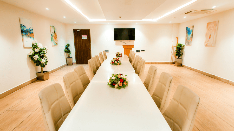 A wide-angle shot of a boardroom at Ilara Hotels & Spa, Tada, with a long white conference table surrounded by cushioned chairs facing a wall-mounted television at the far end of the room.
