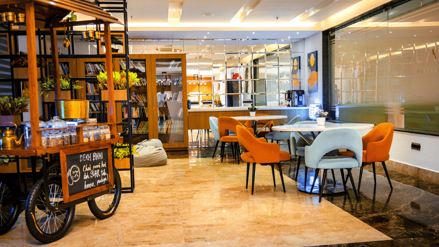 Lobby lounge with a tea cart, plant shelves, book cabinets and round tables with orange and mint chairs at Ilara Hotels & Spa, Tada.