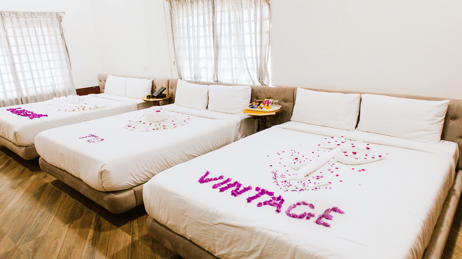 A large bedroom featuring 3 beds with white linens on a wooden floor, with pink petals on the closest bed spelling the word VINTAGE | Vintage Trails By Ilara