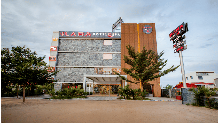 Angled view of the facade of Ilara Hotels & Spa, Tada, with a wood-panelled tower, rooftop signage, palm trees and cars along the entrance drive.