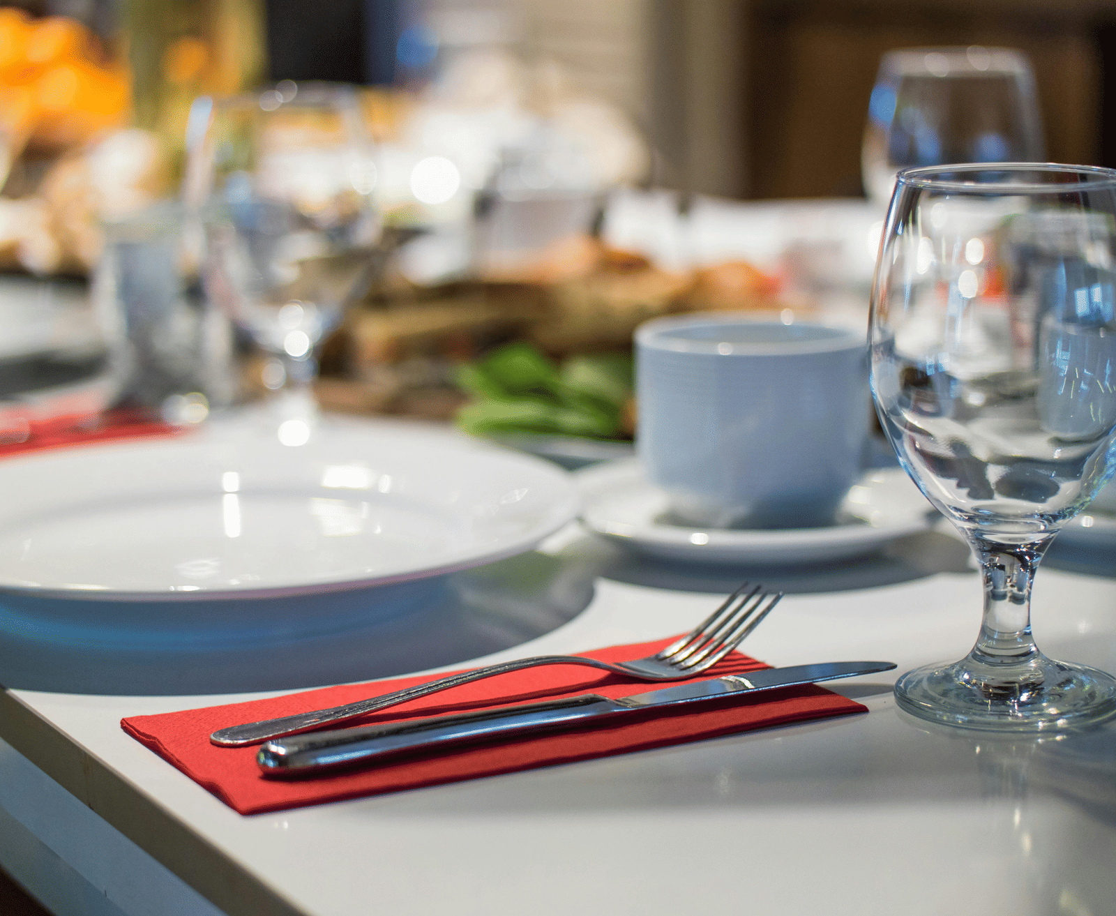 A close-up, blurred shot of a formal dinner table setting with white plates, red napkins, silverware, and clear wine glasses.