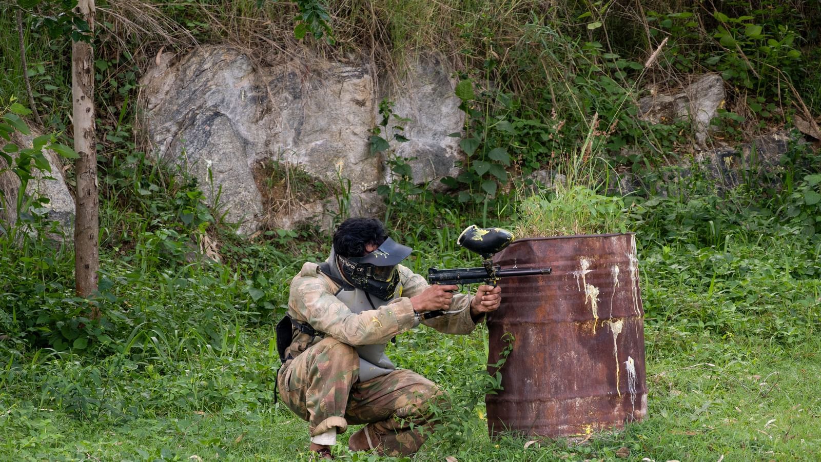 Two individuals are engaged in a paintball game in a natural setting, with one person taking cover behind a wooden barrier | RD's Nature Retreat, Bangalore