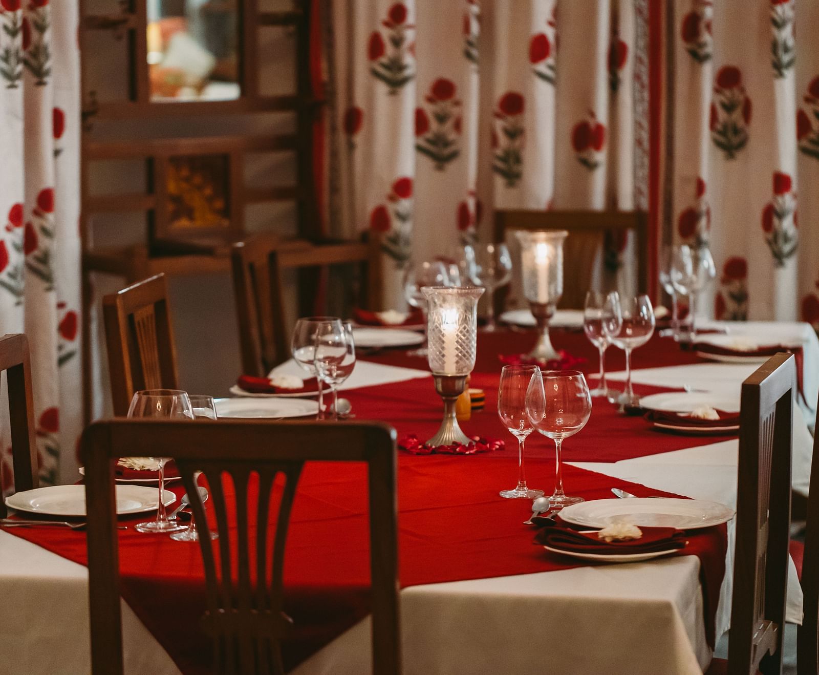 A dining table set with plates, glasses, and candles, surrounded by chairs and patterned curtains- Shahpura Bagh. 