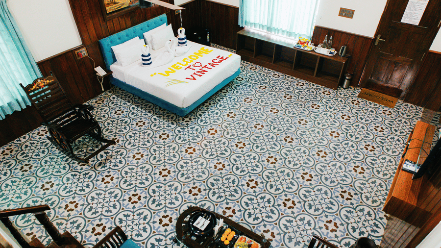 A high-angle view of a bedroom featuring patterned floor tiles, a double bed with a blue headboard, and seating furniture | Vintage Trails By Ilara