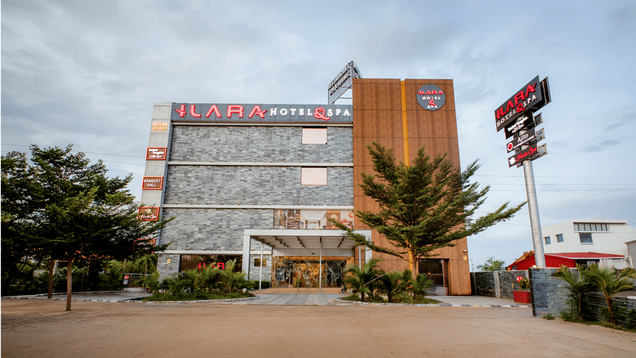 Angled view of the facade of Ilara Hotels & Spa, Tada, with a wood-panelled tower, rooftop signage, palm trees and cars along the entrance drive.
