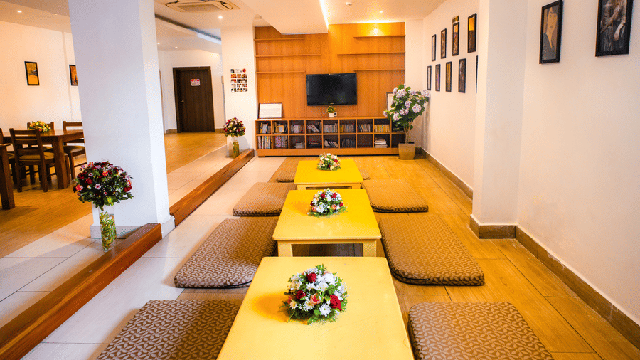 Japanese-style floor seating with low tables and cushions beside book shelves and a television in a wooden shelf wall at Ajito, Ilara Hotels & Spa, Tada.