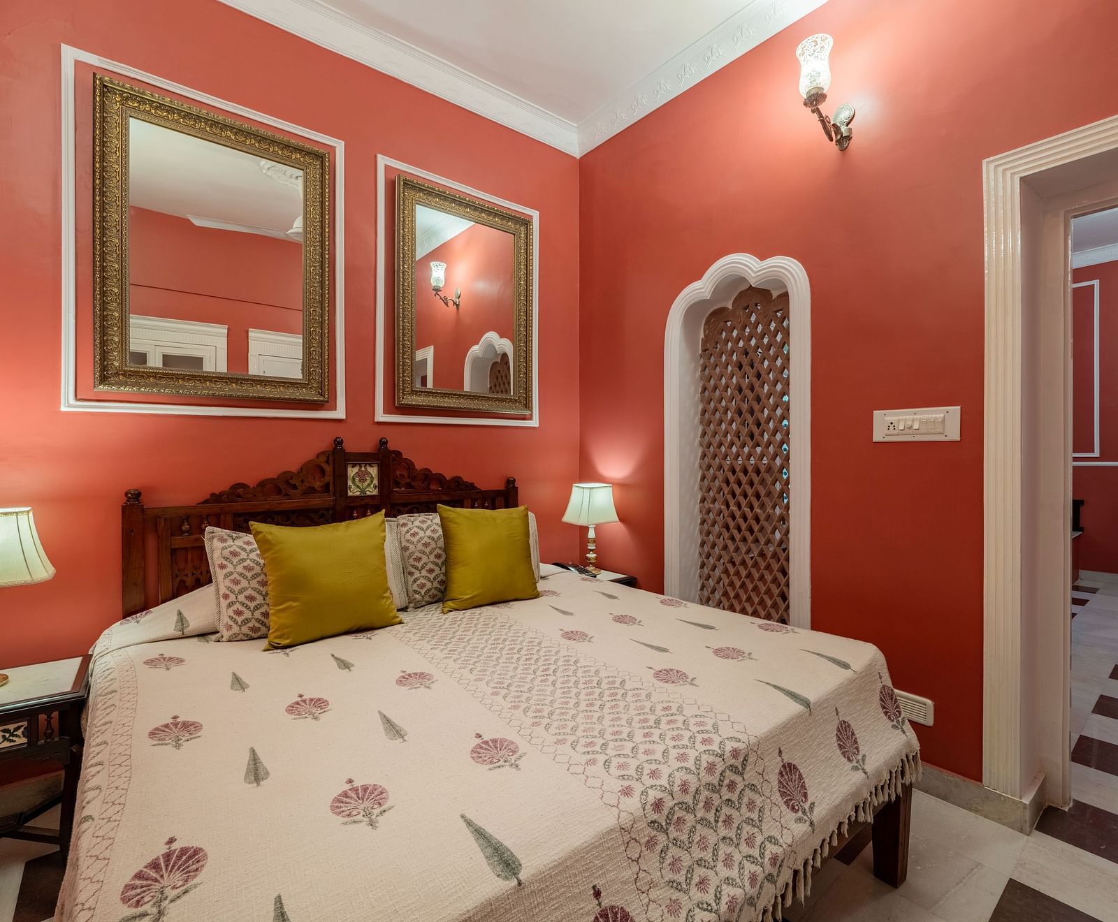 A vibrant bedroom at Dera Rawatsar, Jaipur, features a comfortable bed, framed mirrors, bedside lamps, and an ornate niche in the wall.