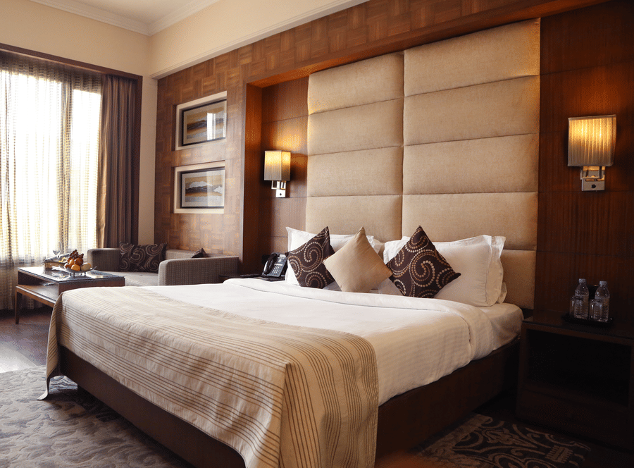Premium Rooms at The Bristol Hotel Gurgaon, with a large bed, spreads, pillows, lamps, a window, a table and a cushion.