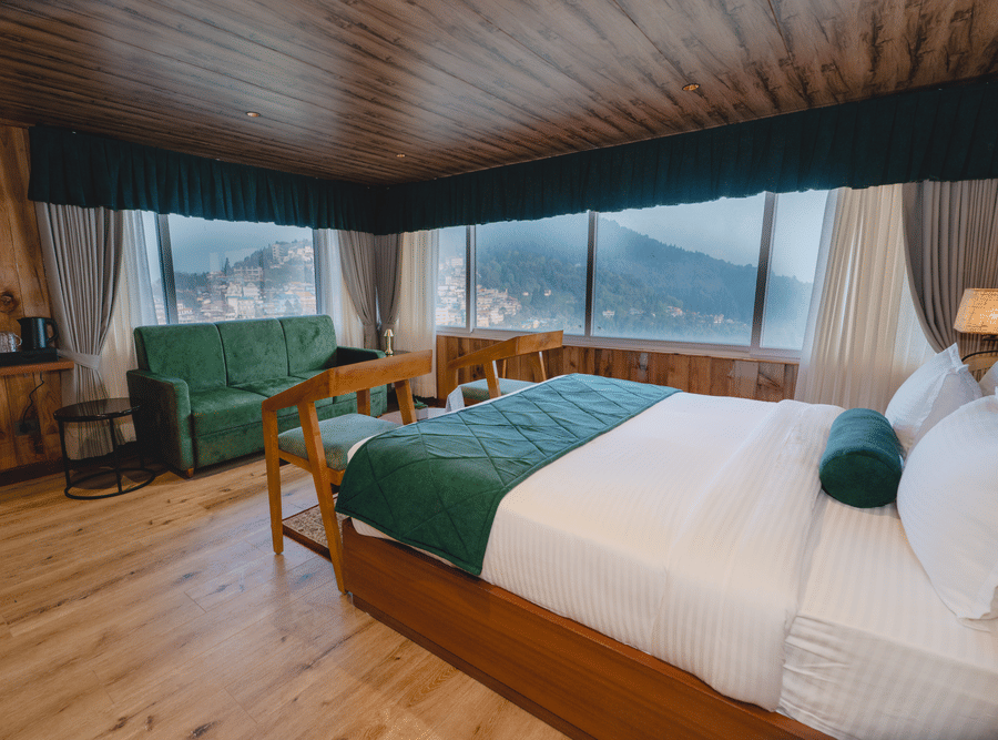 Spacious hotel room with wooden interiors king size bed green sofa seating and large windows offering scenic hillside views | Mount Lungta Boutique Hotel in Darjeeling