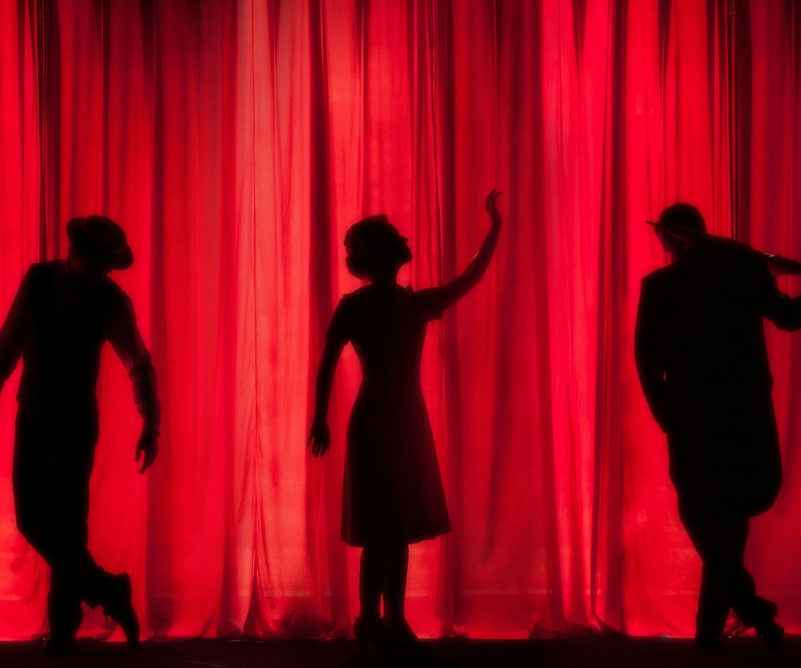 Three actors' silhouettes seen on a theatre stage with a red curtain behind them