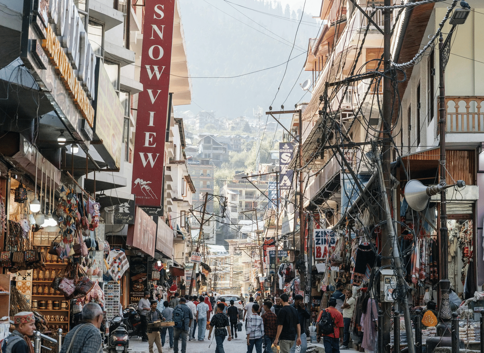 A busy street with shops, hanging signboards, electric poles, and people walking between buildings in a market area with hills in the background.