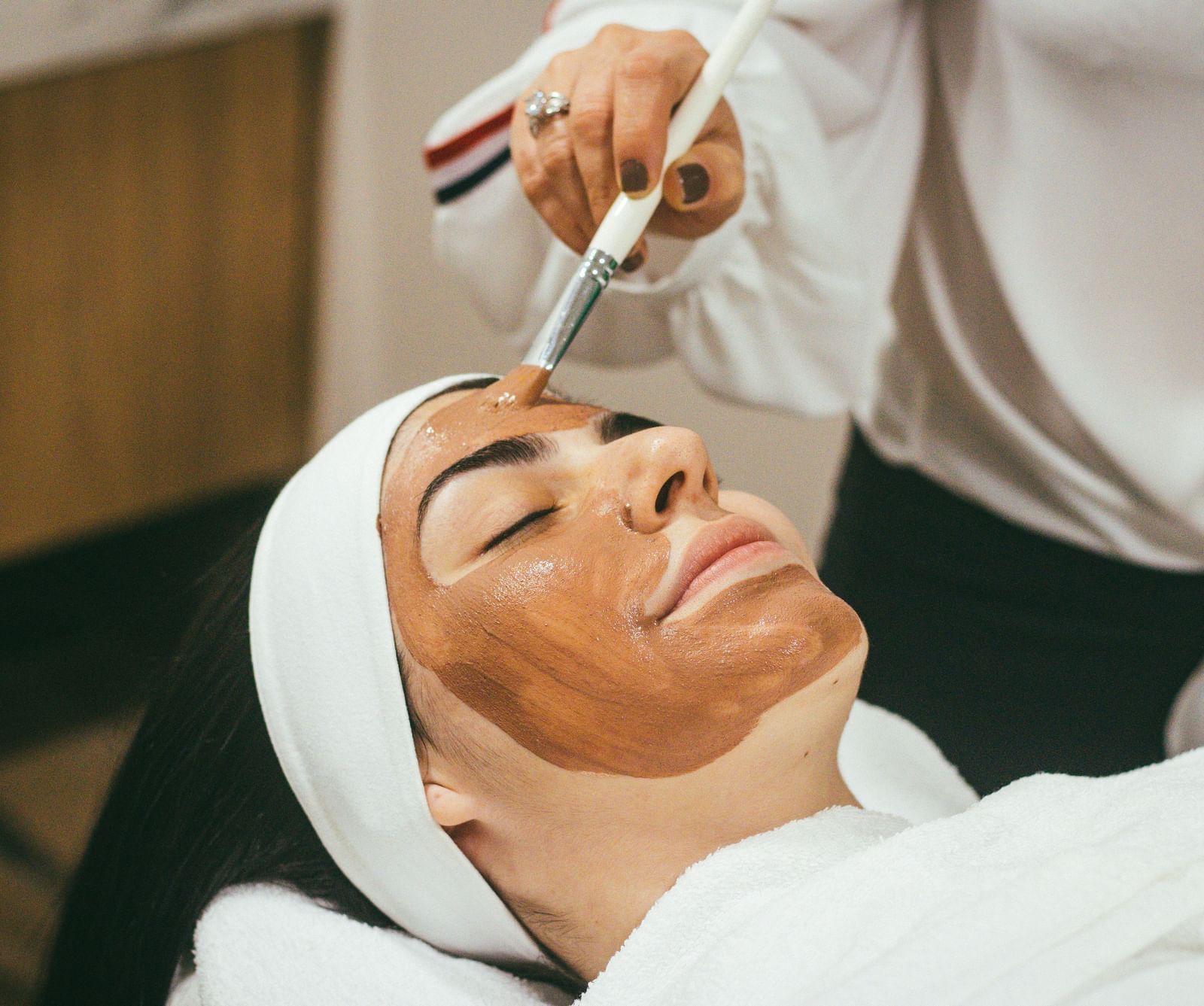 A woman lying down with her eyes closed enjoying a mud mask being applied on her face with the help of a brush.